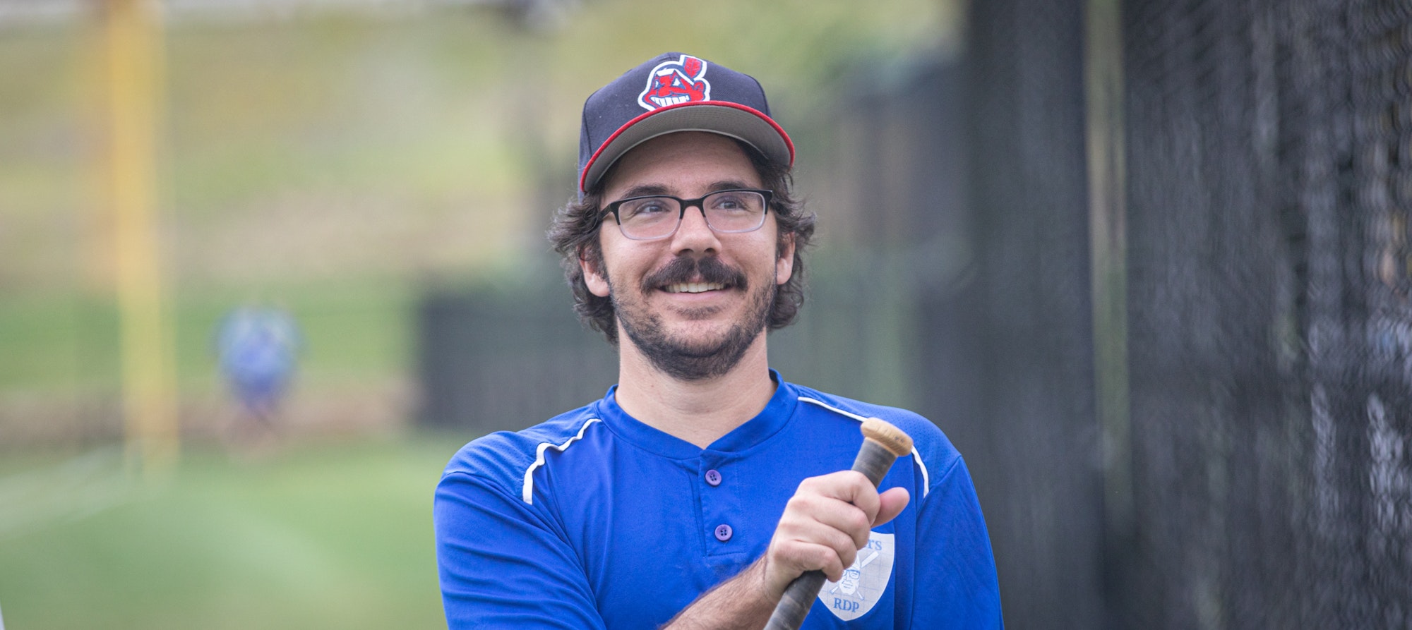 A smiling man in a baseball cap and blue jersey holding a baseball bat.