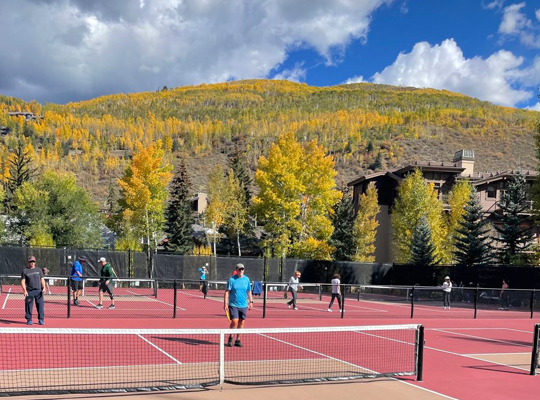 People playing pickleball on outdoor courts with autumn-colored trees and a mountain in the background.