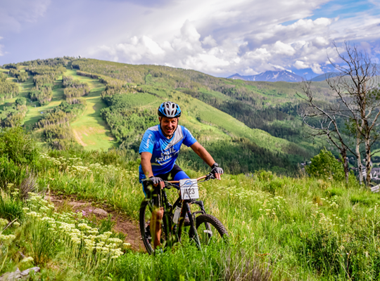 A cyclist on a mountain trail, lush greenery, and hills in the background.
