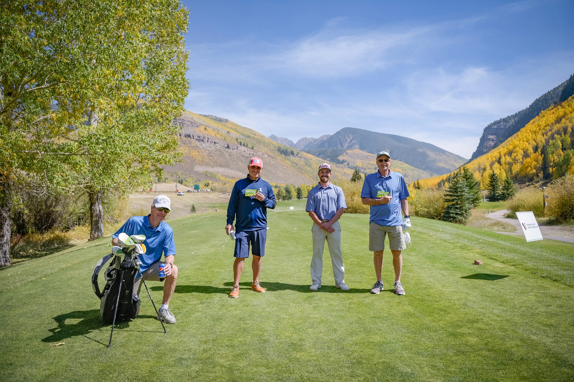 Four men at a golf course with autumn trees and mountains in the background.