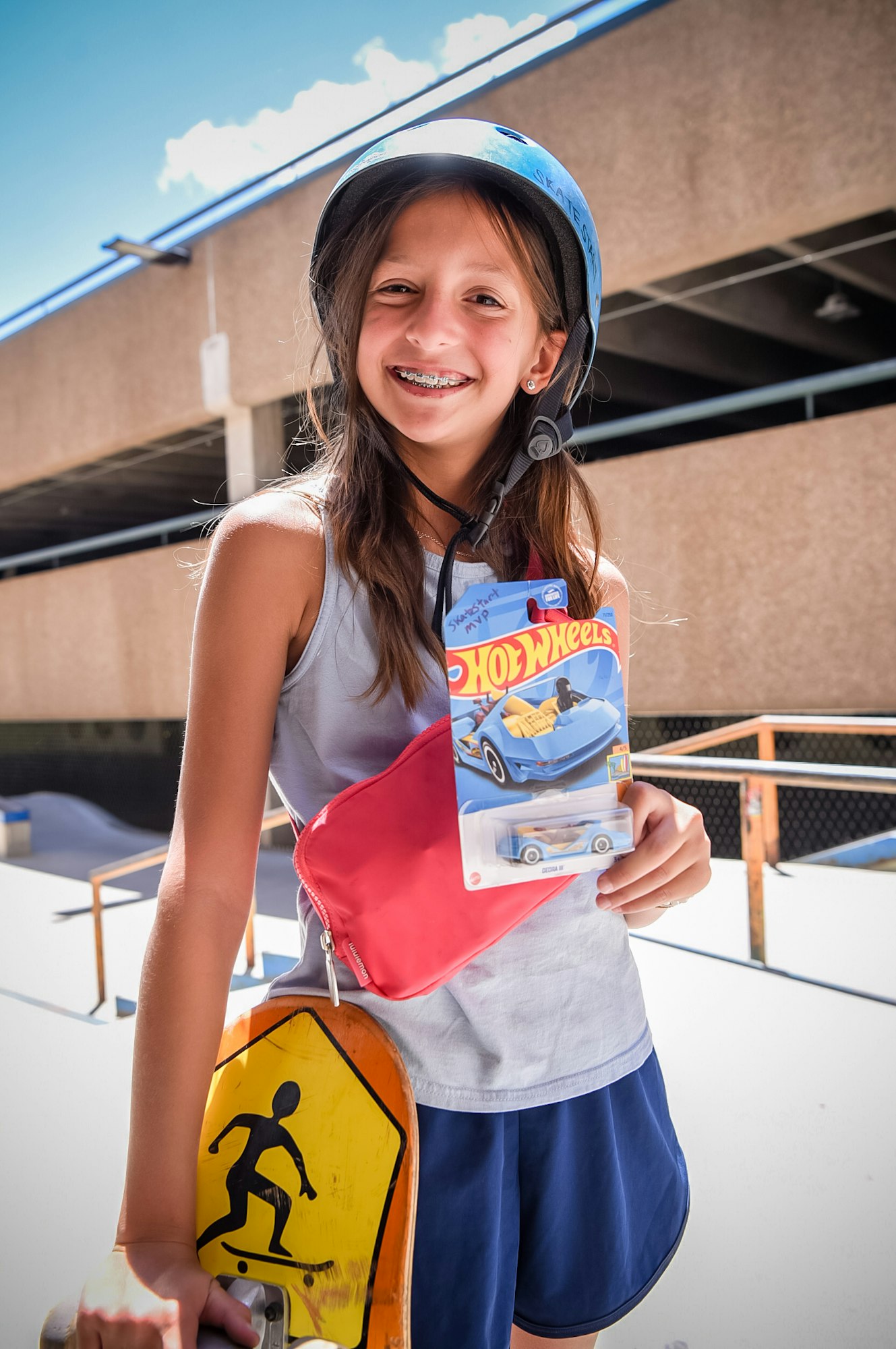 A smiling child with a helmet holding a Hot Wheels toy and a skateboard at a skatepark.