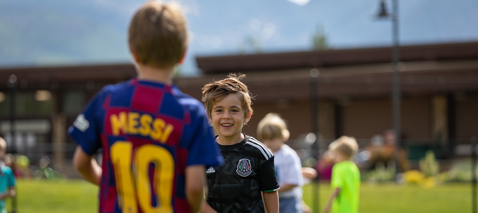 Two kids in soccer jerseys with one wearing a Messi shirt, smiling outside on a sunny day.