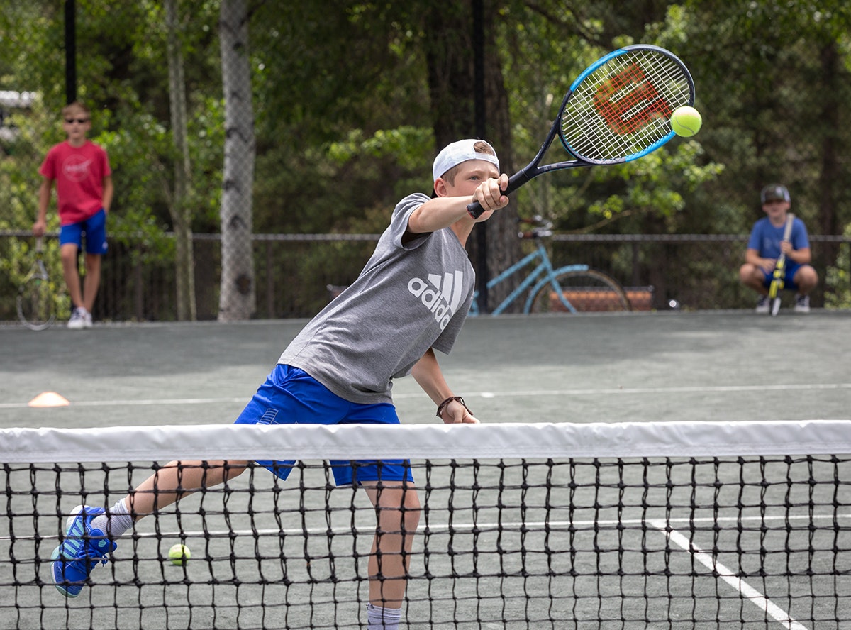 A boy playing tennis, hitting a forehand, with others waiting in the background on a court.