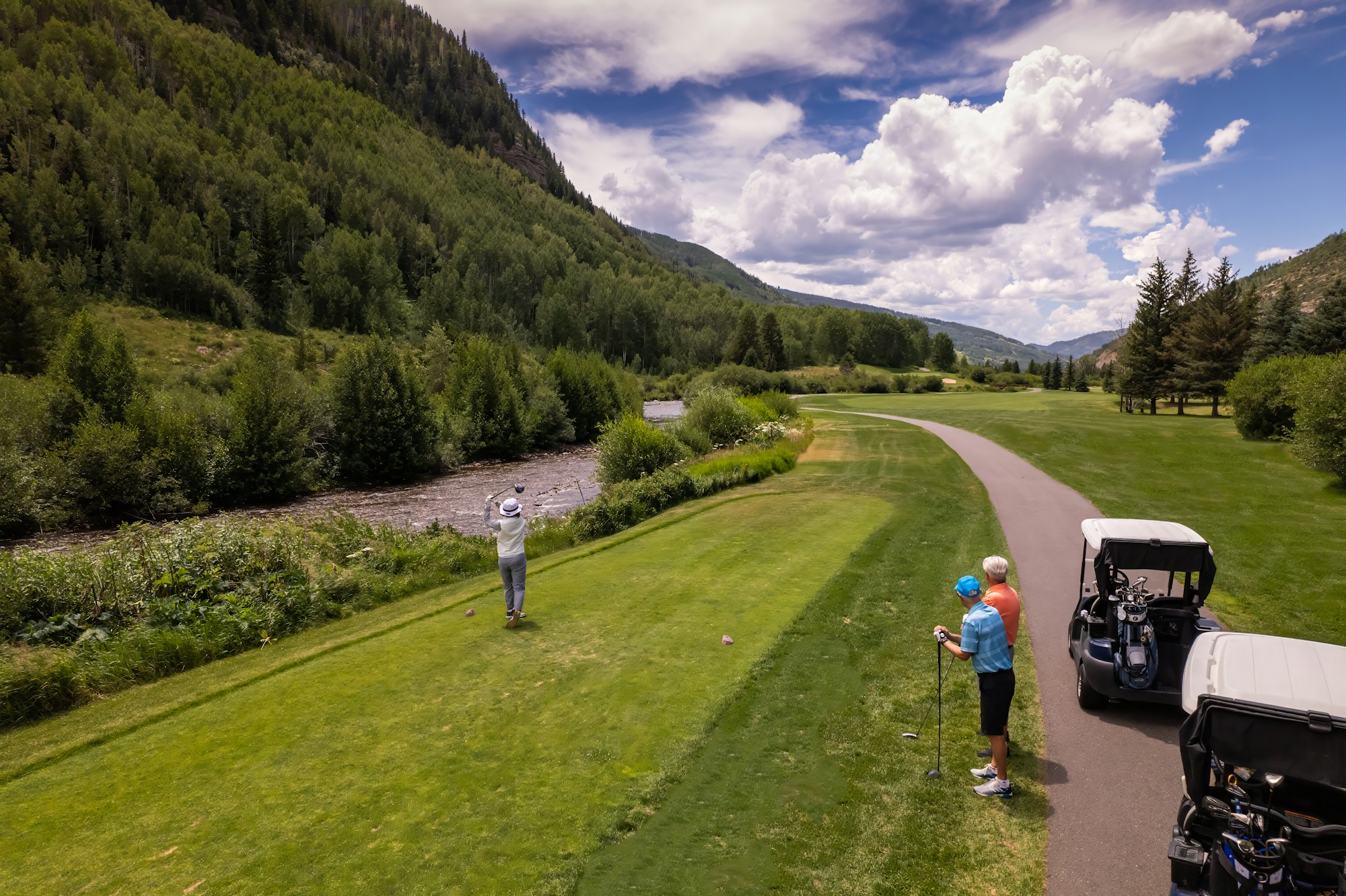 A scenic golf course with players, a golf cart, green trees, and a cloudy blue sky.