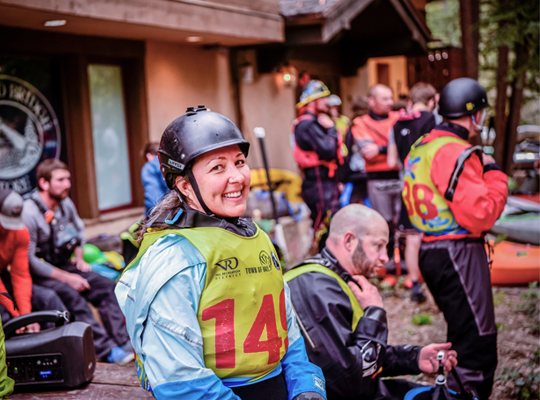A person in a helmet and life vest smiles, surrounded by others with kayaking gear.