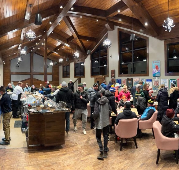 A bustling indoor gathering with people conversing and seated at tables, under a wooden beamed ceiling with hanging lights.