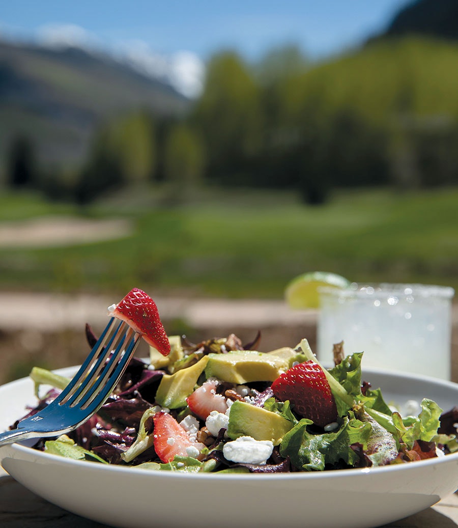 A fresh salad with strawberries, avocado, greens, and cheese, a fork, and a blurry natural backdrop.