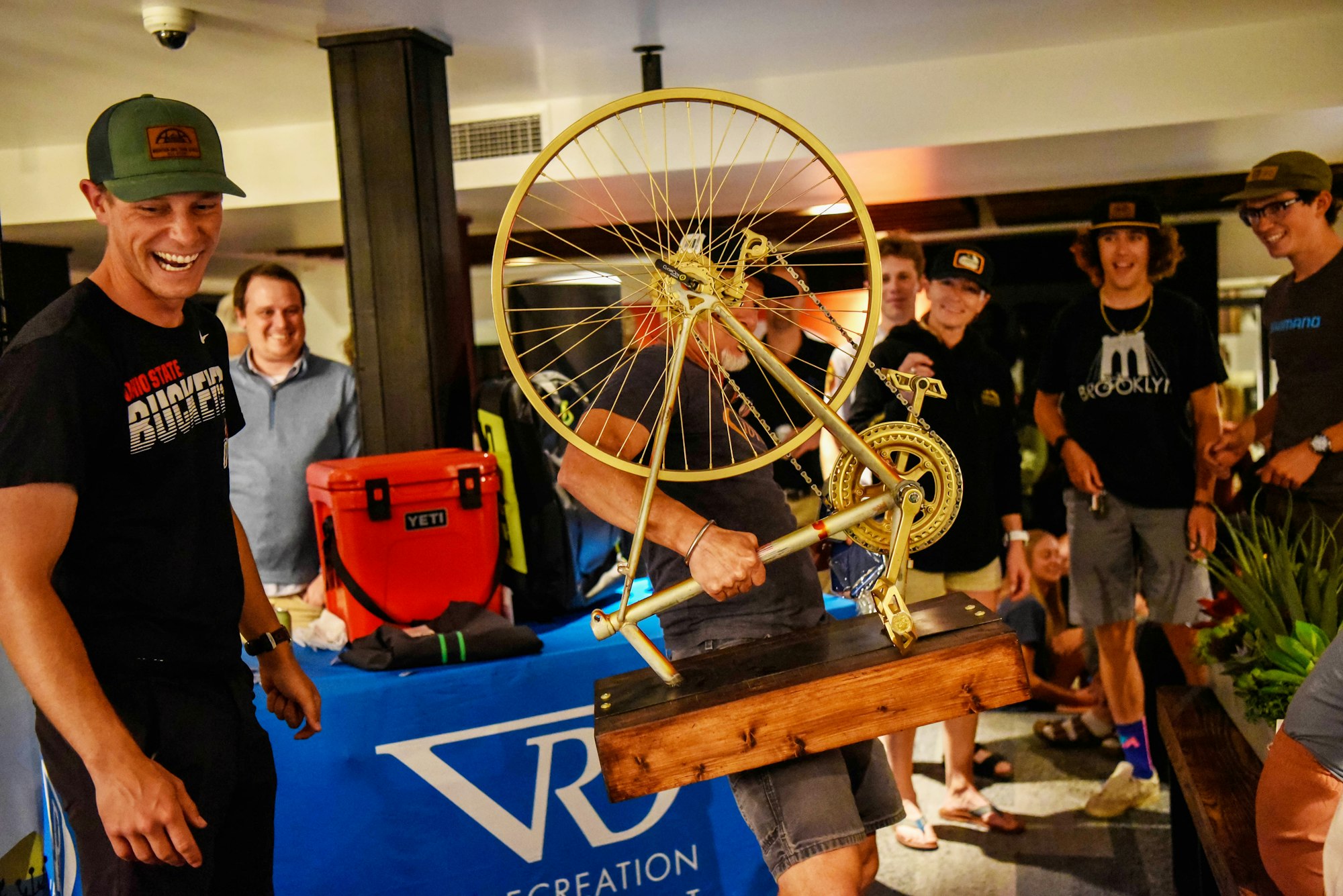 Group of people enjoying a trophy with a bicycle wheel, sprocket, and pedal mounted on wood.