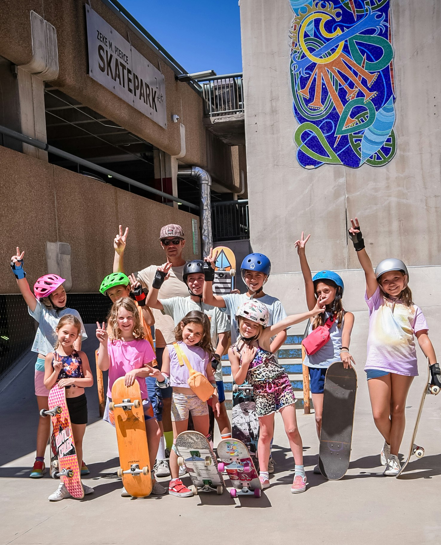 A group of kids and an adult with skateboards, wearing helmets, at a skatepark, smiling and posing for the photo.