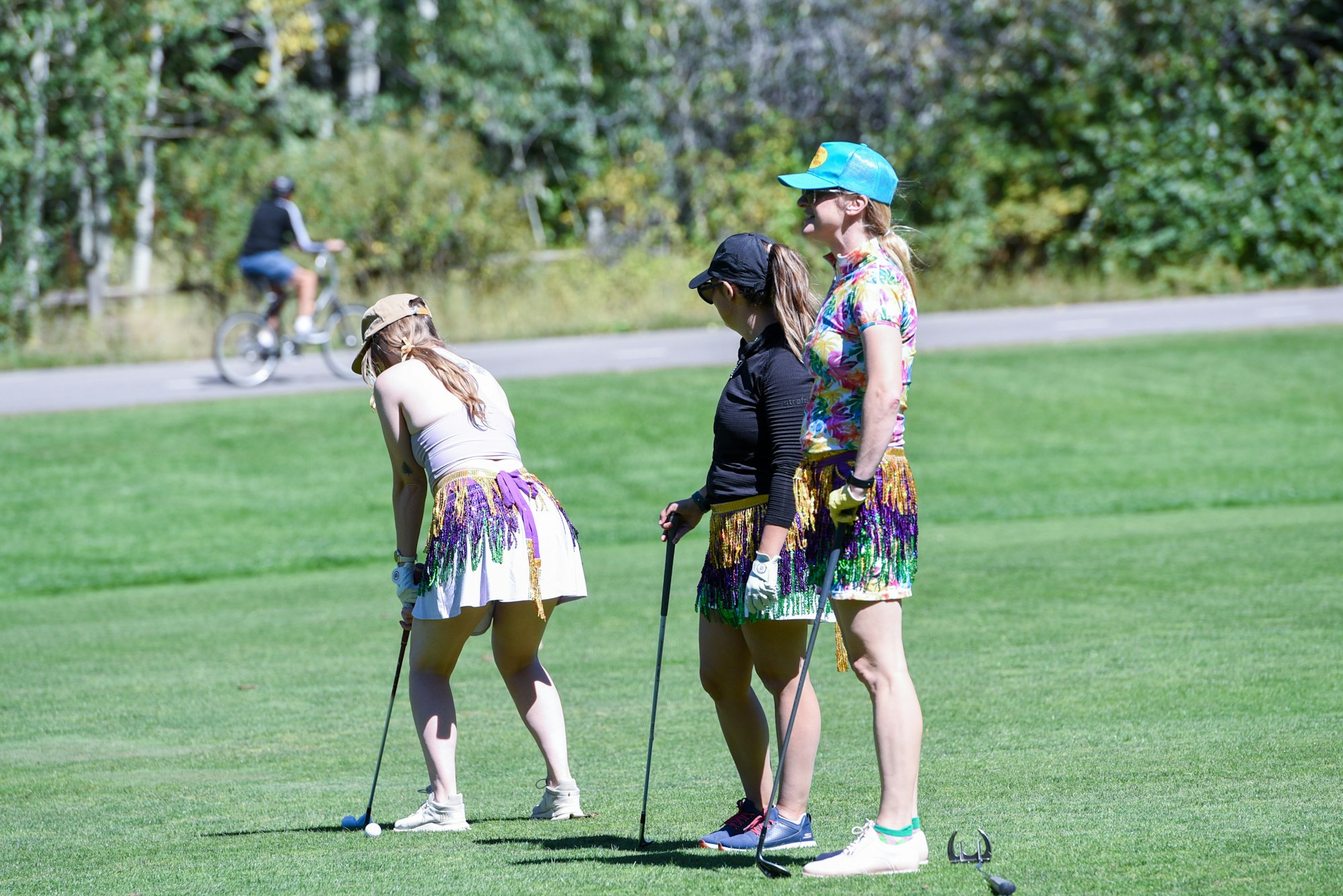 Three women in colorful outfits playing golf on a green, with a cyclist in the background.