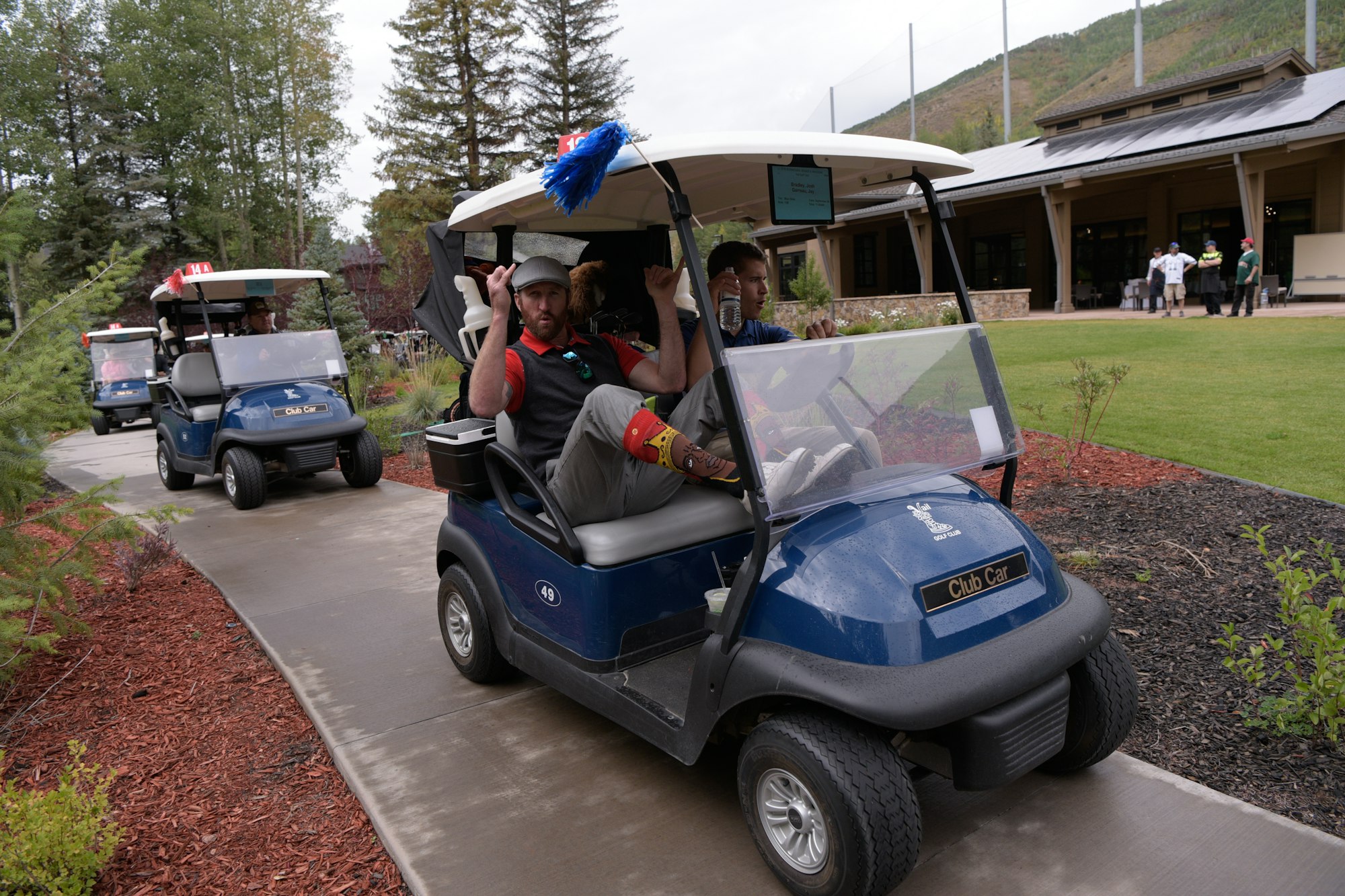 Two men in a golf cart with decorative feathers, another cart behind, near a building with people.