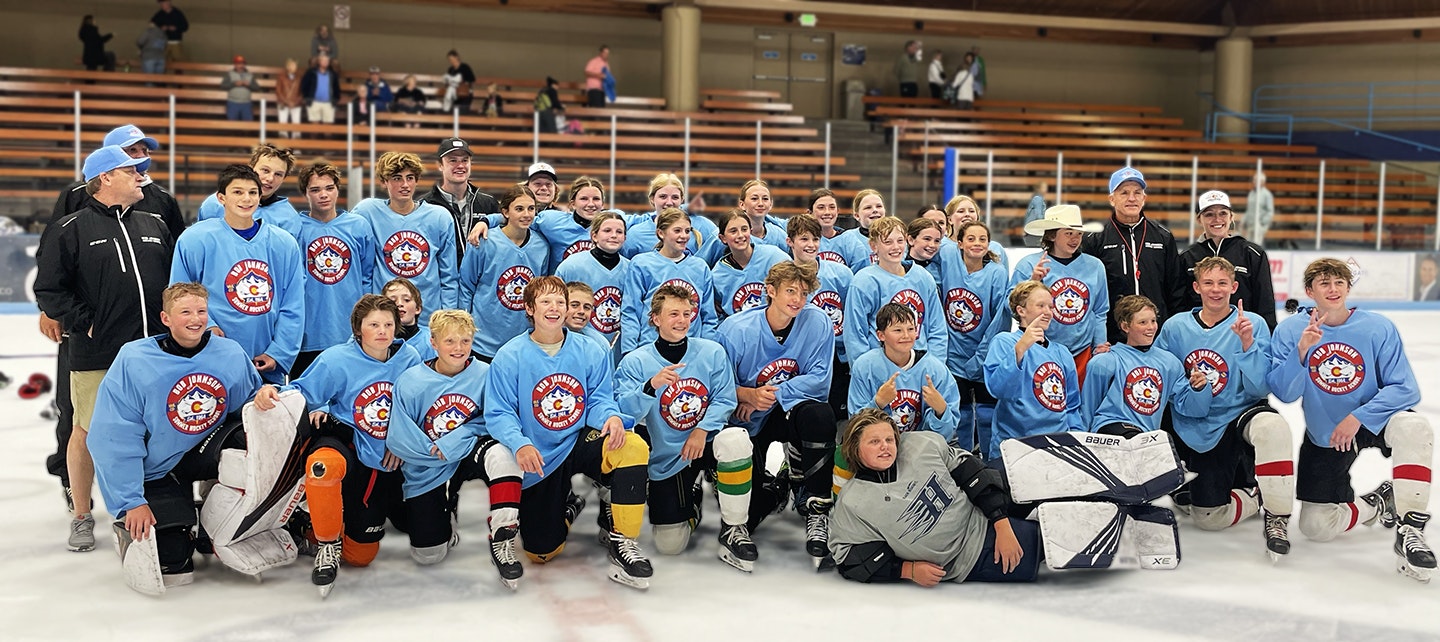 Group of youth ice hockey players and coaches posed on ice, smiling, wearing matching blue sweatshirts with a team logo.