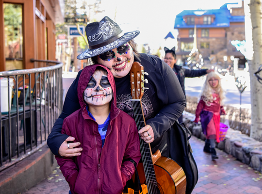 Two people with face paint and a guitar smiling, possibly at a festive or Halloween event, with someone in the background.