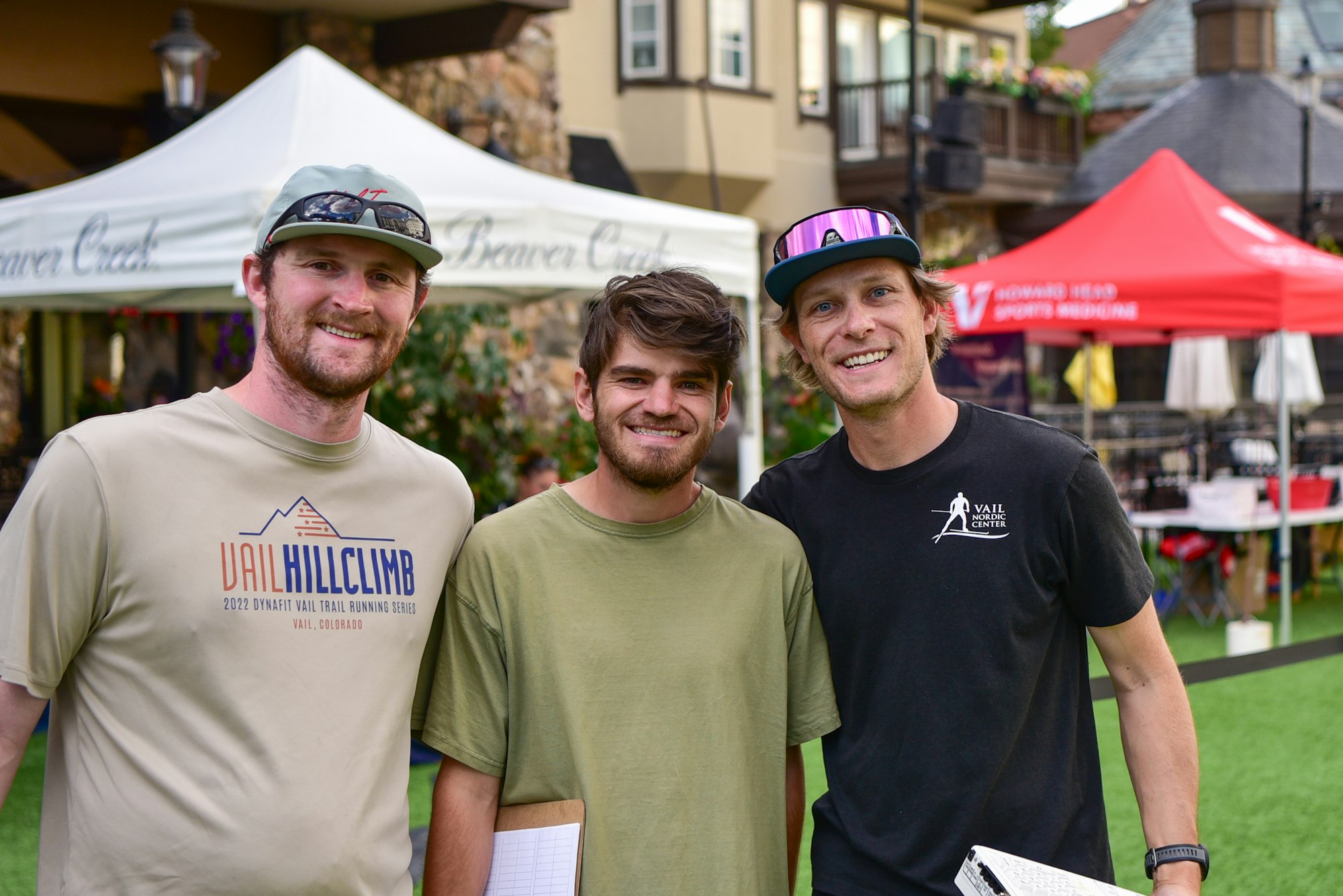 Three people smiling at an outdoor event with tents and a building in the background.