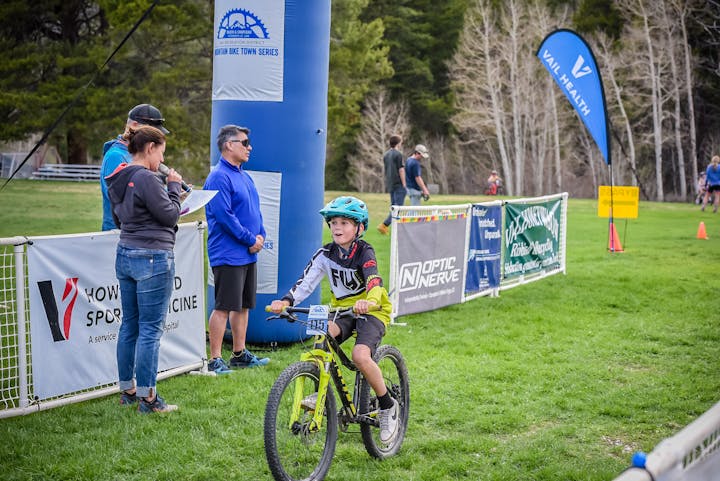A child mountain biker at a race finish line with onlookers and event banners.
