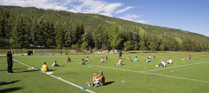 Outdoor children’s sports training session on a green field with a forest backdrop under a clear sky.