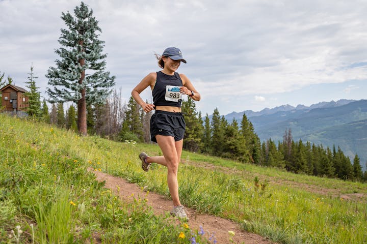 A person is trail running on a scenic mountain path, surrounded by trees and distant hills.