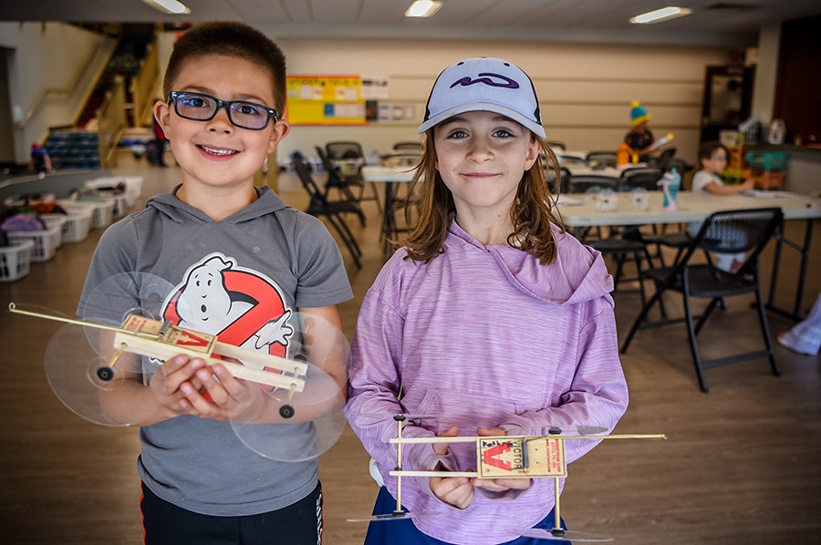 Two kids smiling, each holding a handmade model airplane, in a room with tables and chairs in the background.