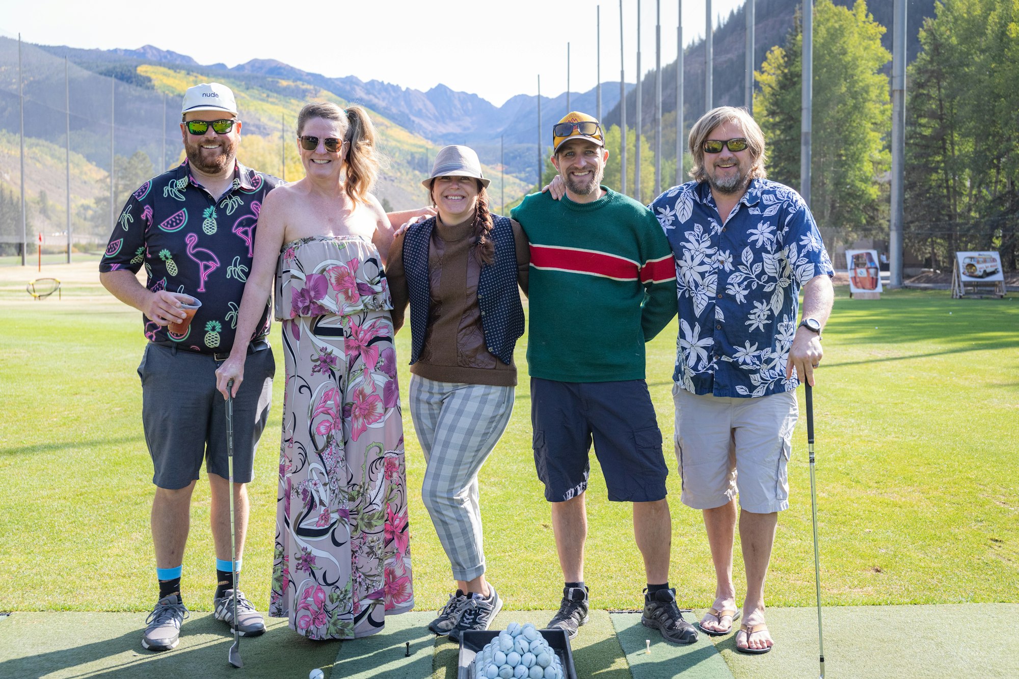 Five people posing with golf clubs on a sunny golf range, dressed in casual, colorful attire.