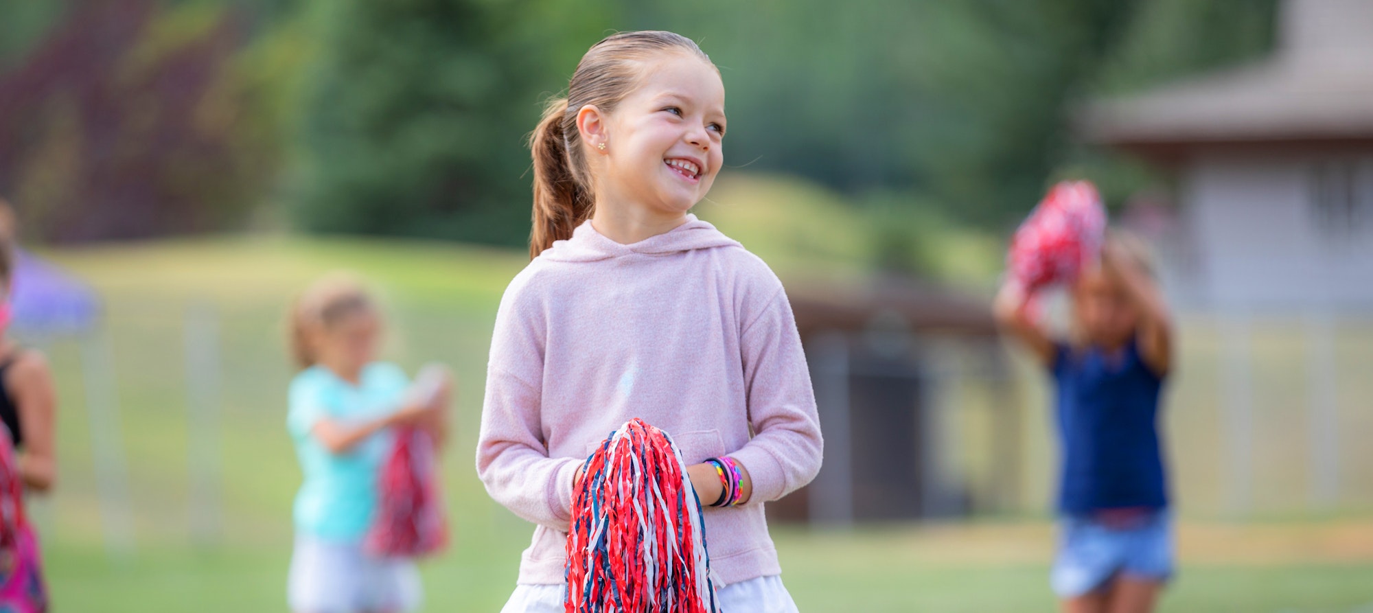 A smiling child holding pom-poms with blurred figures in the background.