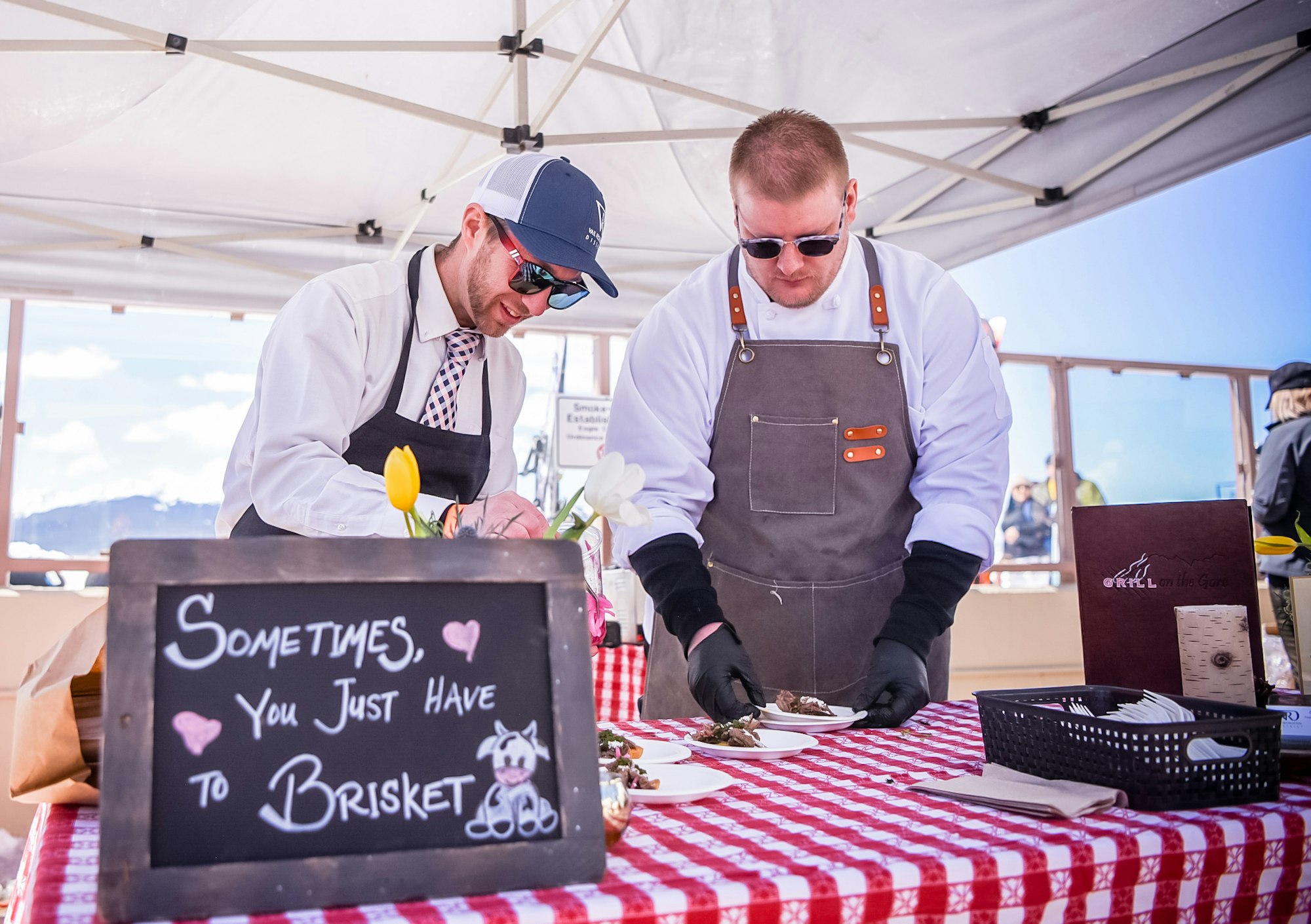 Two chefs preparing food at a brisket-themed outdoor event, with a sign reading "Sometimes, You Just Have to Brisket."
