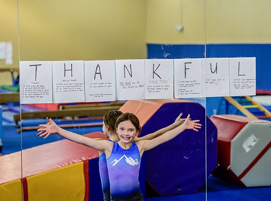 A smiling child in a gymnastics leotard is spreading her arms wide under a sign spelling "THANKFUL" with handwritten notes on it.