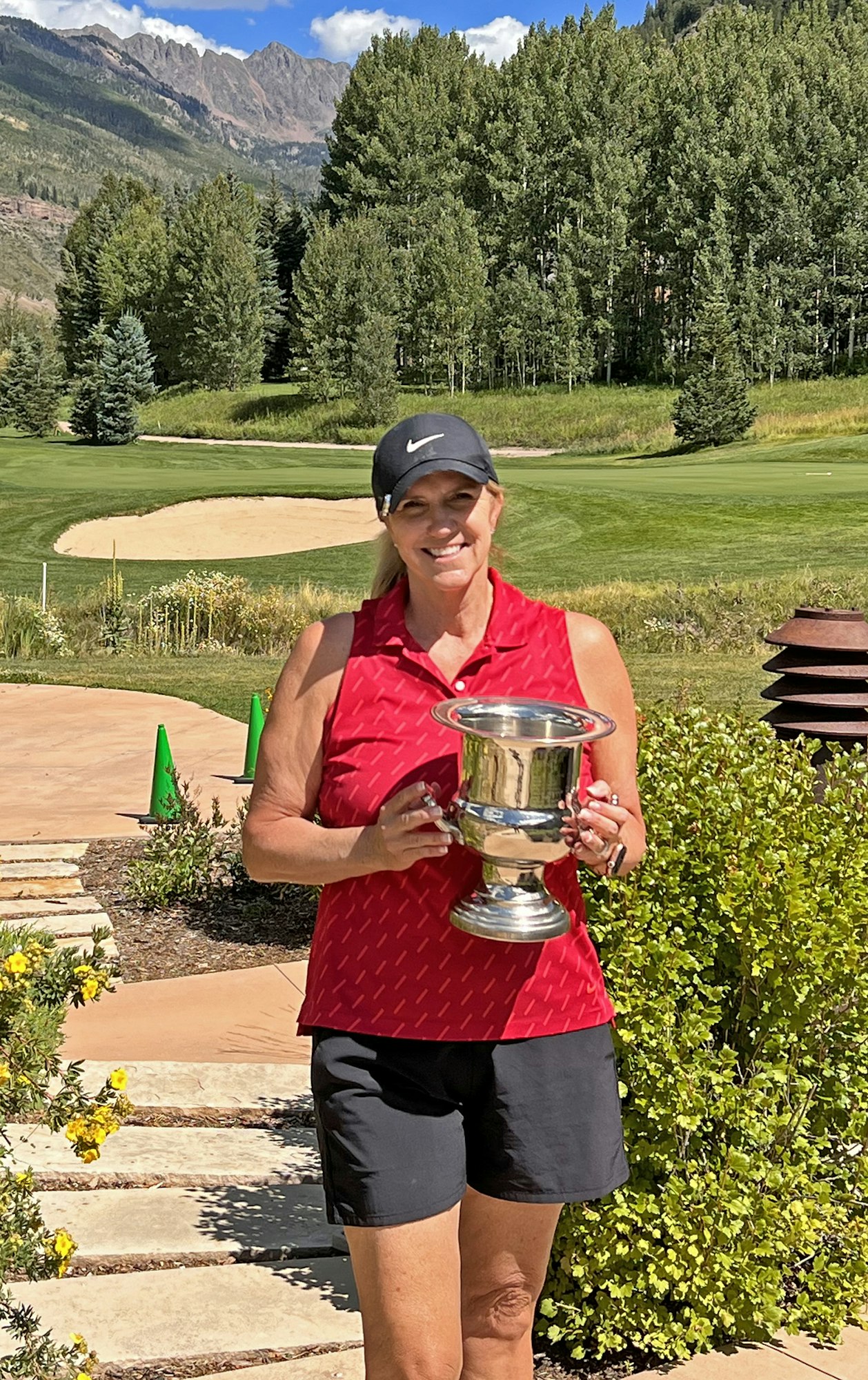 A smiling person holding a trophy on a golf course with mountains in the background.