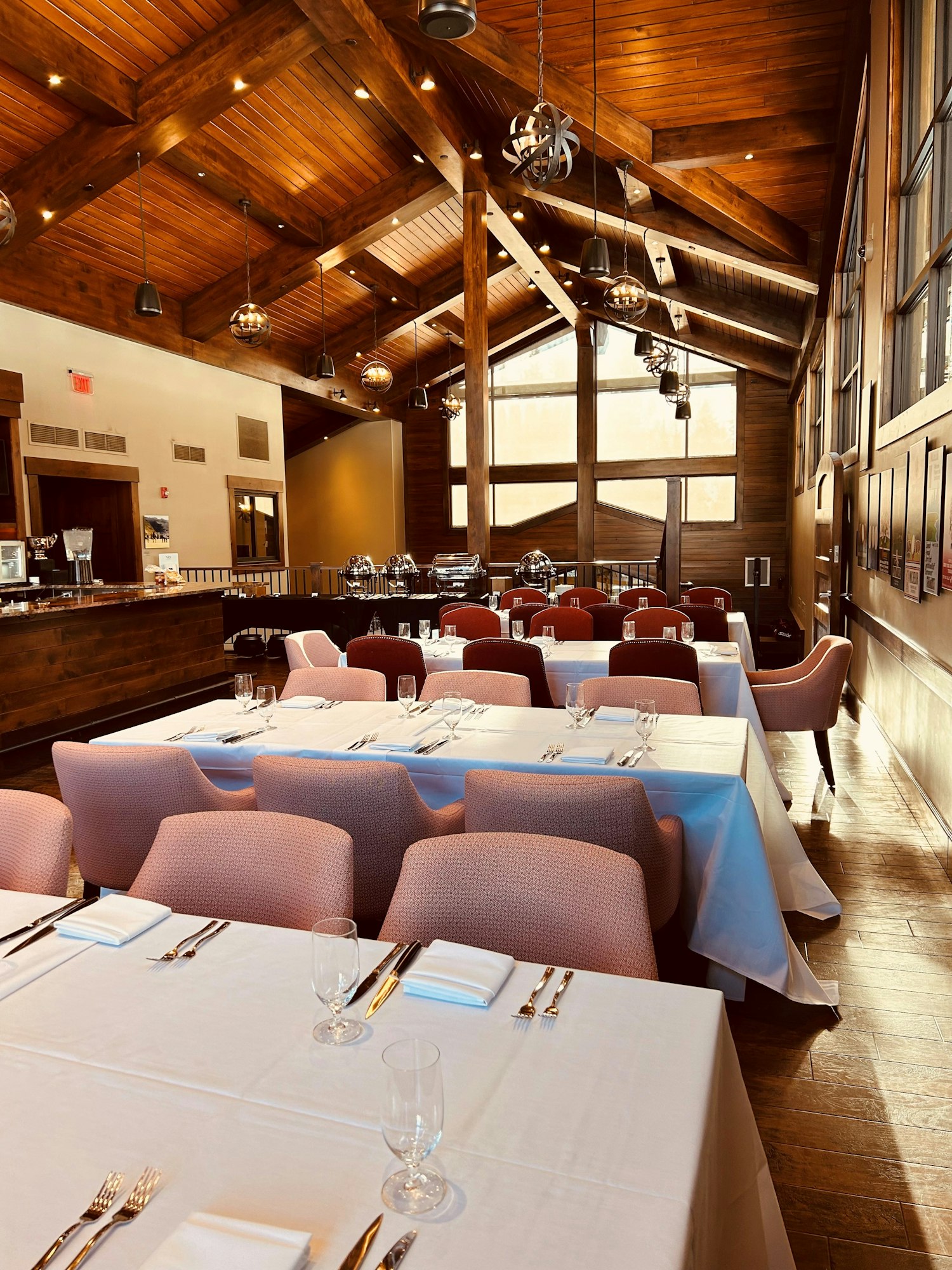 Elegant dining area with wooden ceiling, pink chairs, white tablecloths, and set tables, featuring modern lighting and large windows.
