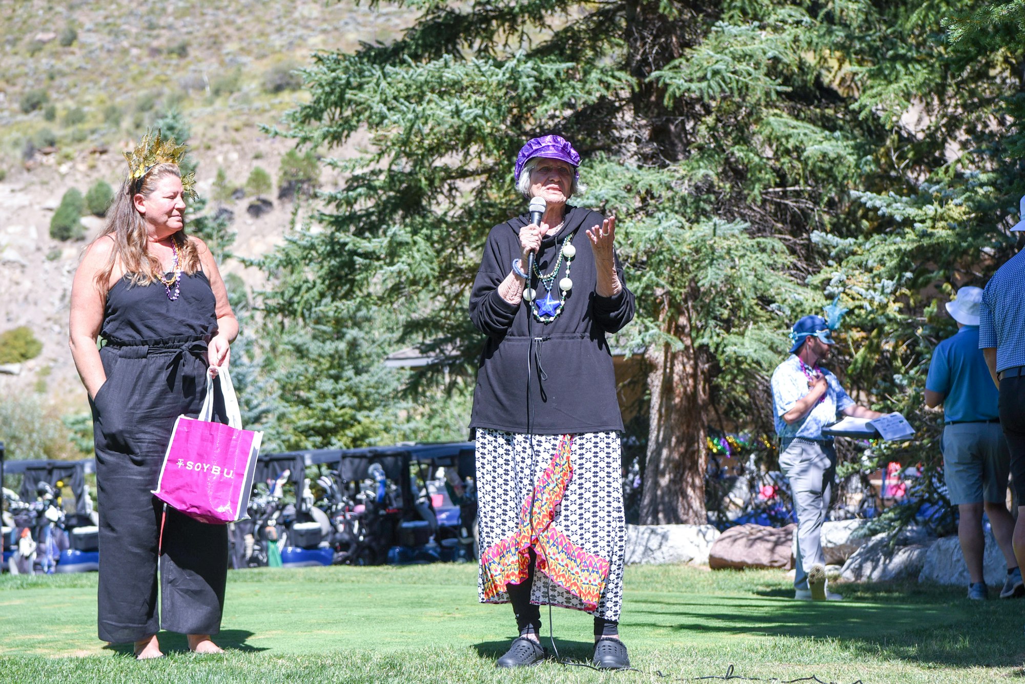 Two people are standing outdoors. One holds a microphone, speaking, while the other carries a pink bag. Trees are in the background.