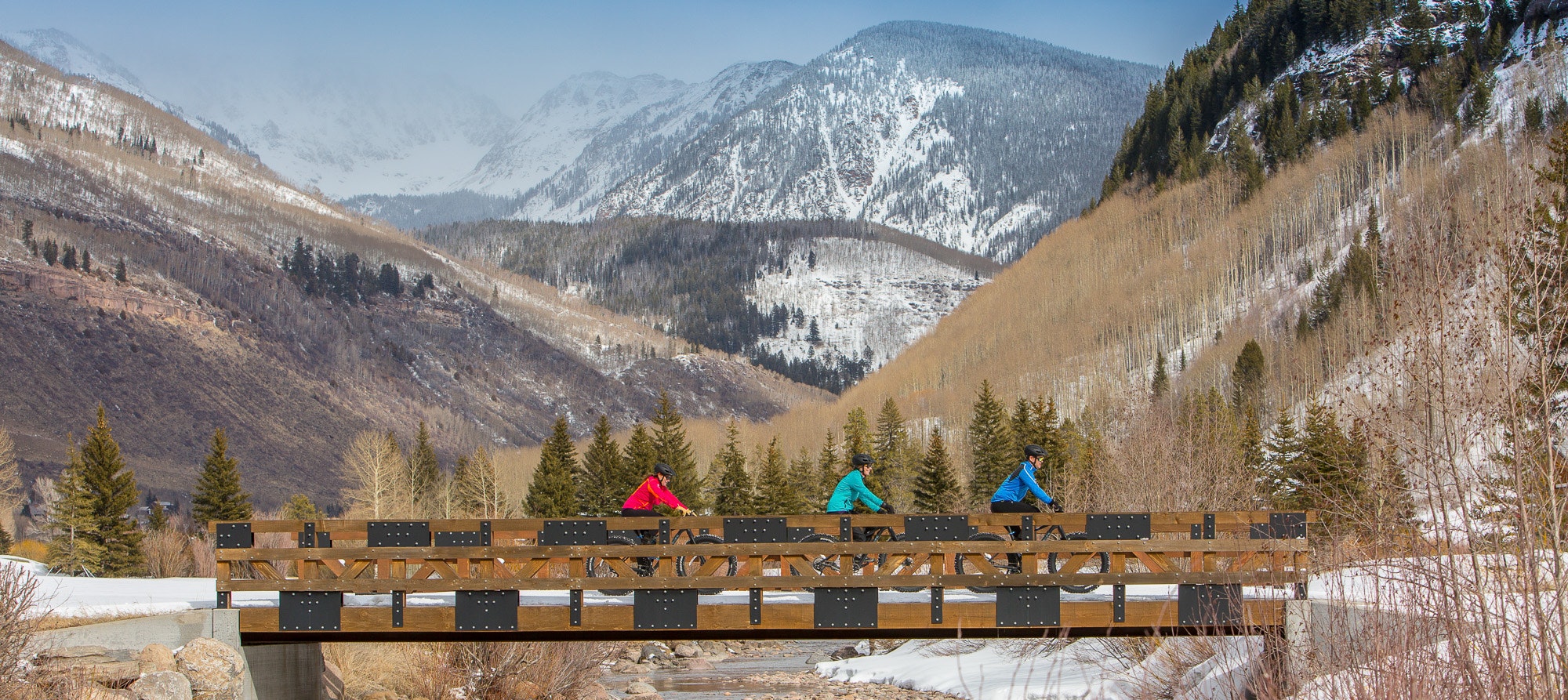 Three cyclists on a wooden bridge with a snowy mountainous backdrop.
