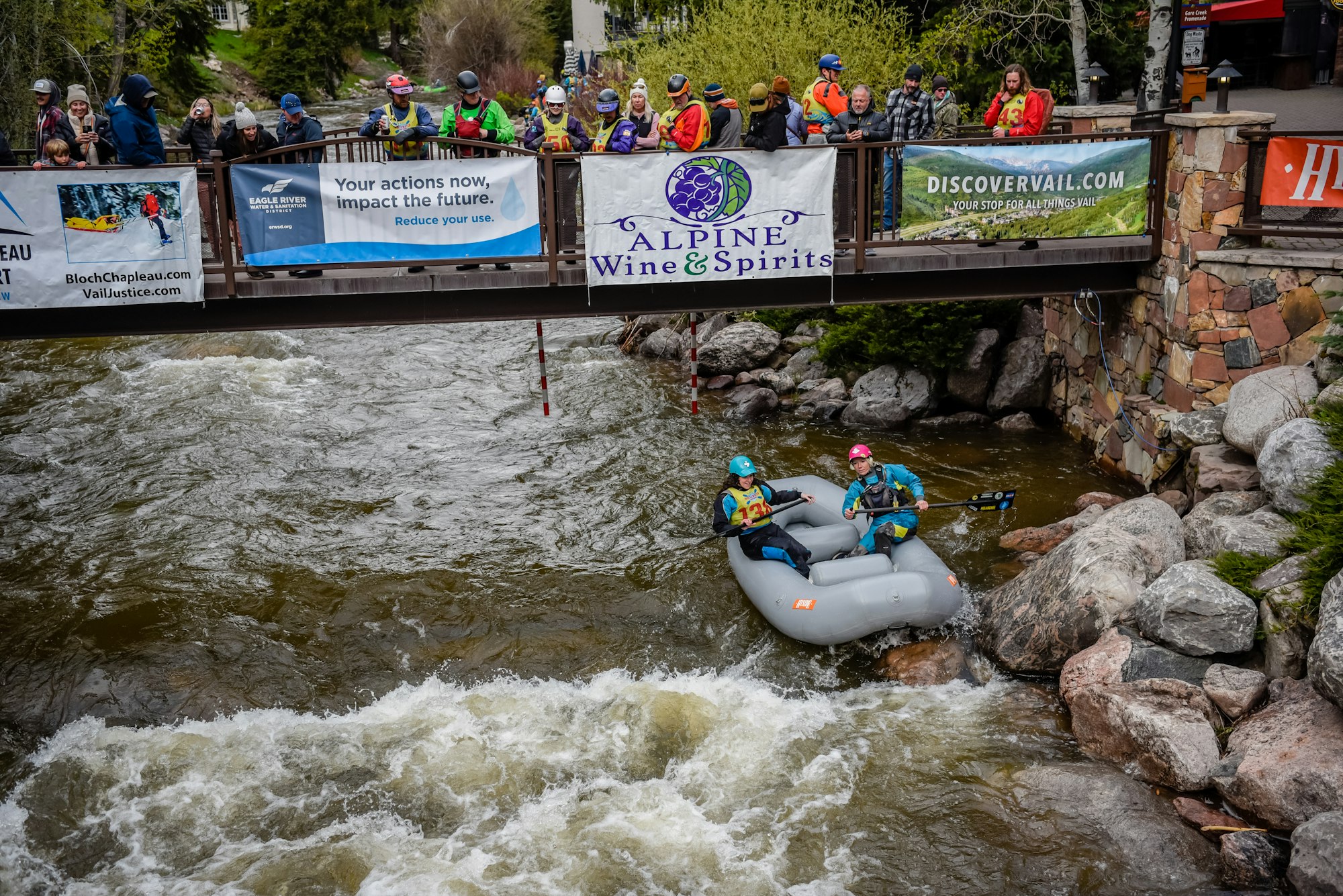 Rafters navigating rough waters, spectators watching from a bridge adorned with banners.