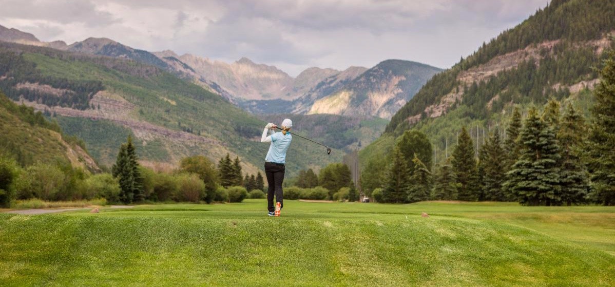 A golfer swinging a club on a course with mountains in the background.