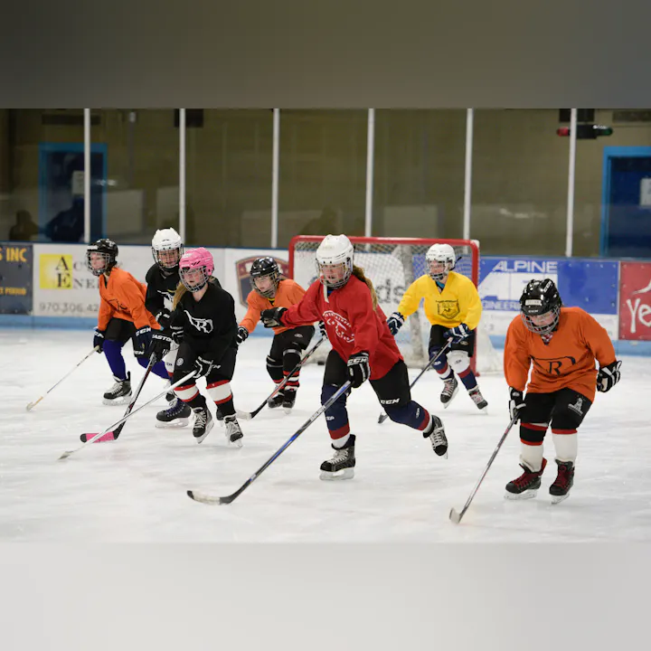 Kids playing ice hockey, wearing colorful jerseys and helmets, holding hockey sticks, on an indoor rink near the goal.