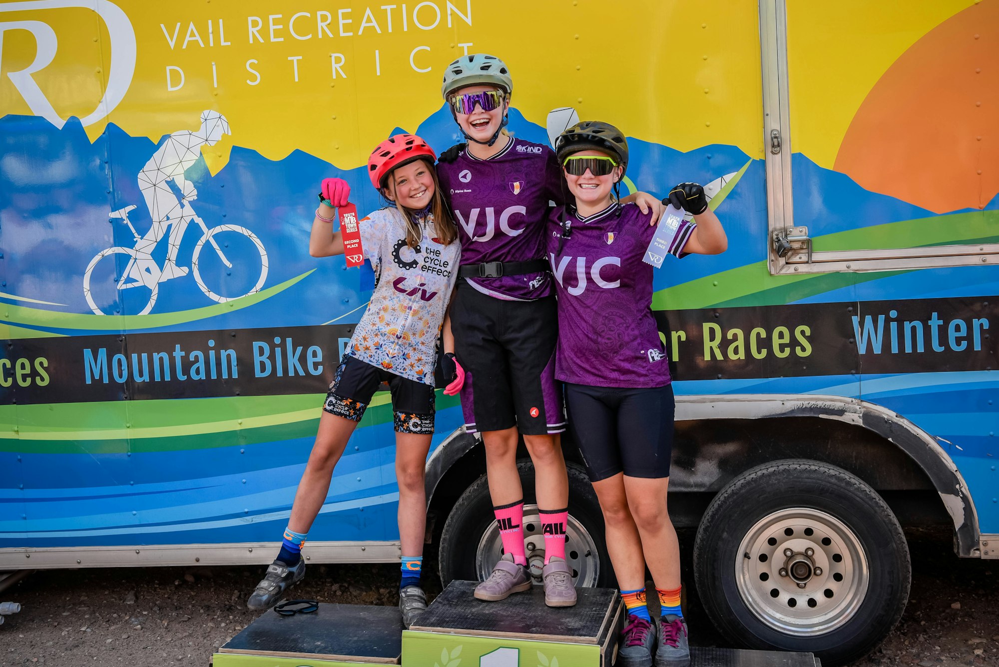 Three happy young cyclists posing with awards in front of a mountain bike race sign.