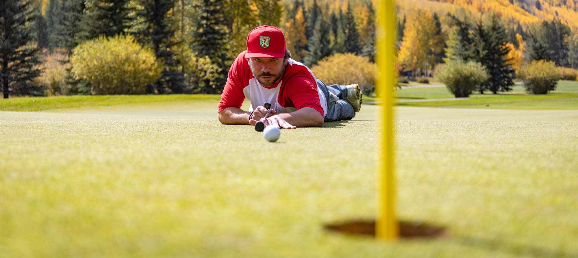 A person lies on a golf course, aligning a putt, with autumn trees in the background.