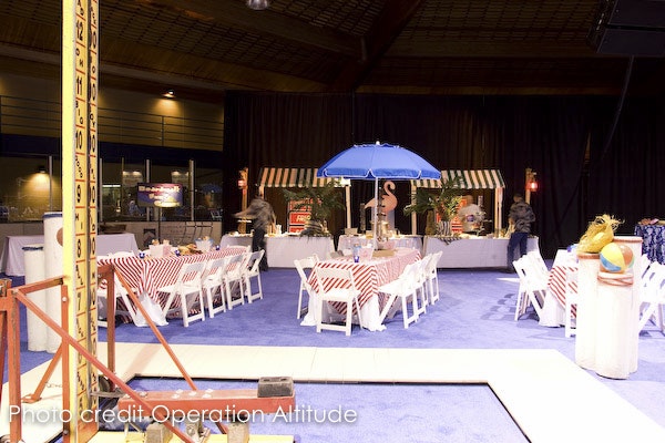 Indoor event space with beach theme, tables with red checkered covers, and a sand area with a miniature golf setup.