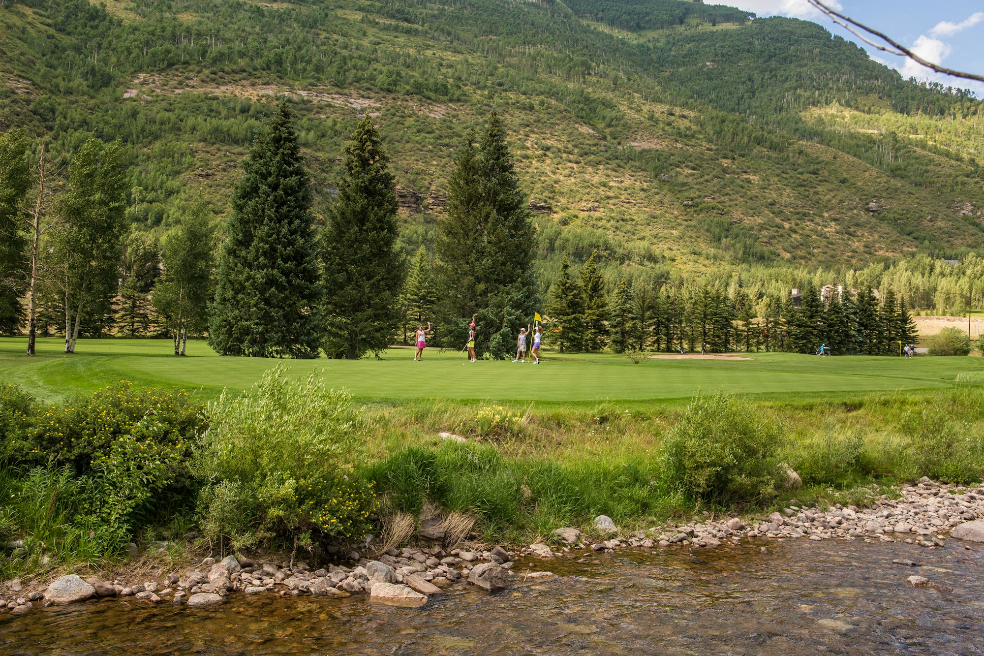 Golf course with players, stream in foreground, wooded hills in the background.