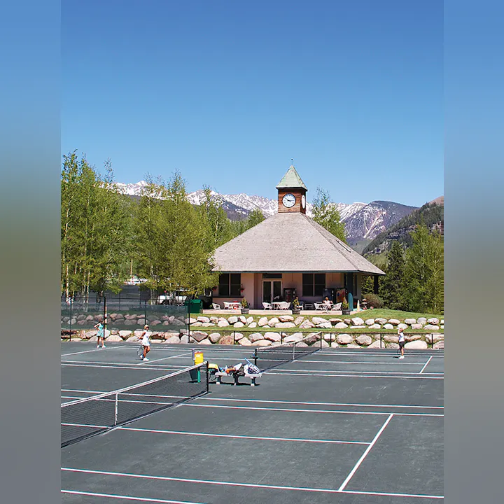 Tennis court with players, a pavilion with a clock tower, and mountains in the background.