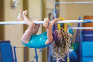 A gymnast hangs upside down from a horizontal bar in a gym, with her legs bent over the bar and arms free.