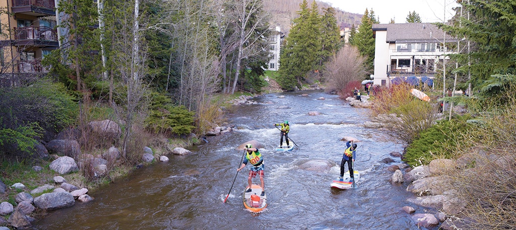 People paddleboarding on a river near forested buildings.