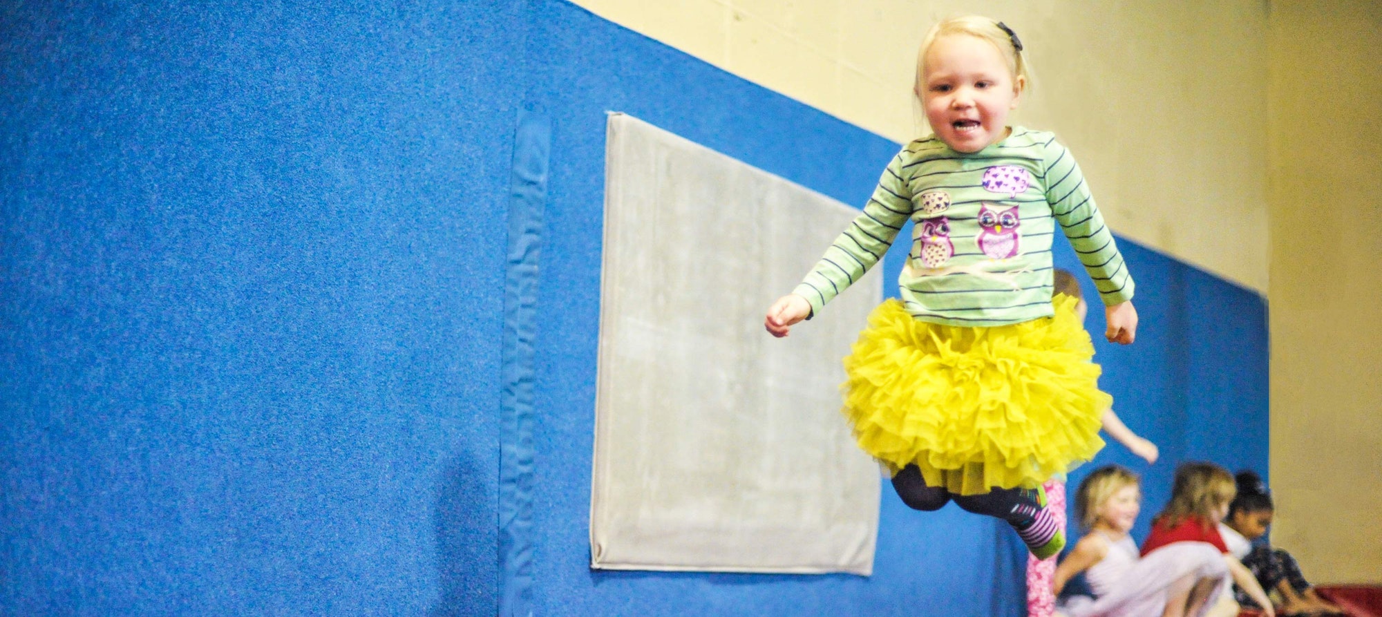 A joyful child in a yellow skirt mid-jump against a blue padded wall with other kids in the background.