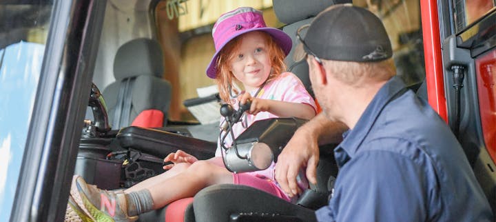 A child in a pink hat smiles at an adult in a vehicle, both hold a steering mechanism.