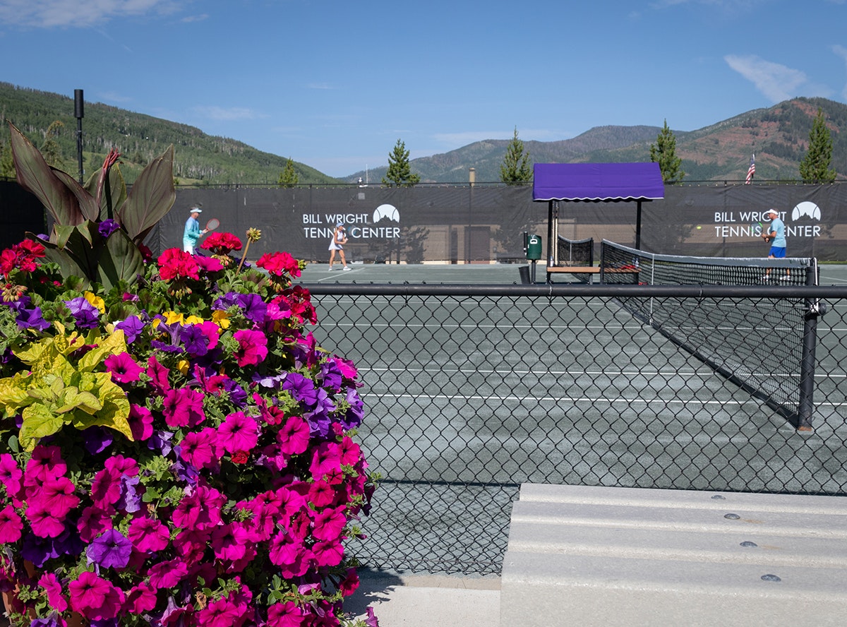 Tennis court with players, colorful flowers in foreground, and mountains in the background.