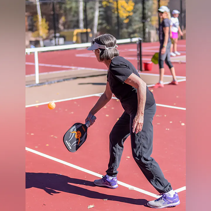 Person plays pickleball on a red court, ready to hit a yellow ball with a paddle.