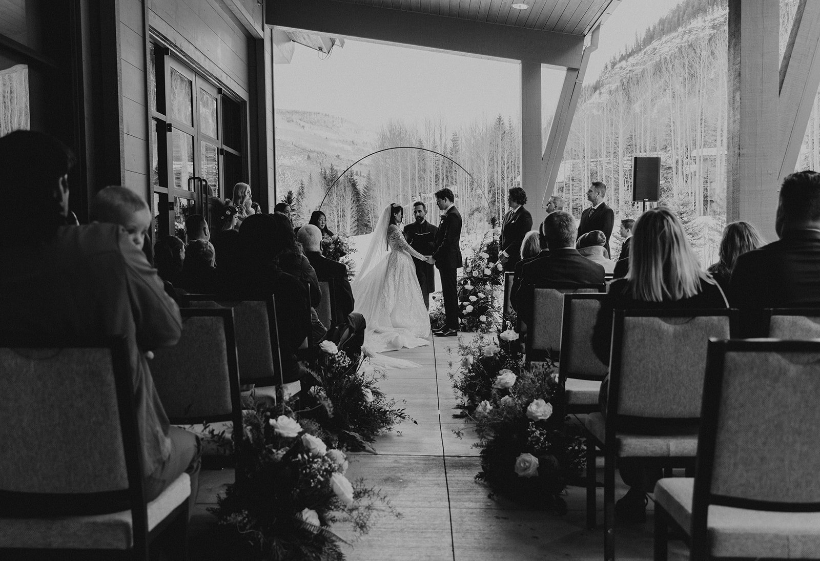 A black-and-white photo of a wedding ceremony with a couple standing outdoors, surrounded by guests and floral arrangements.