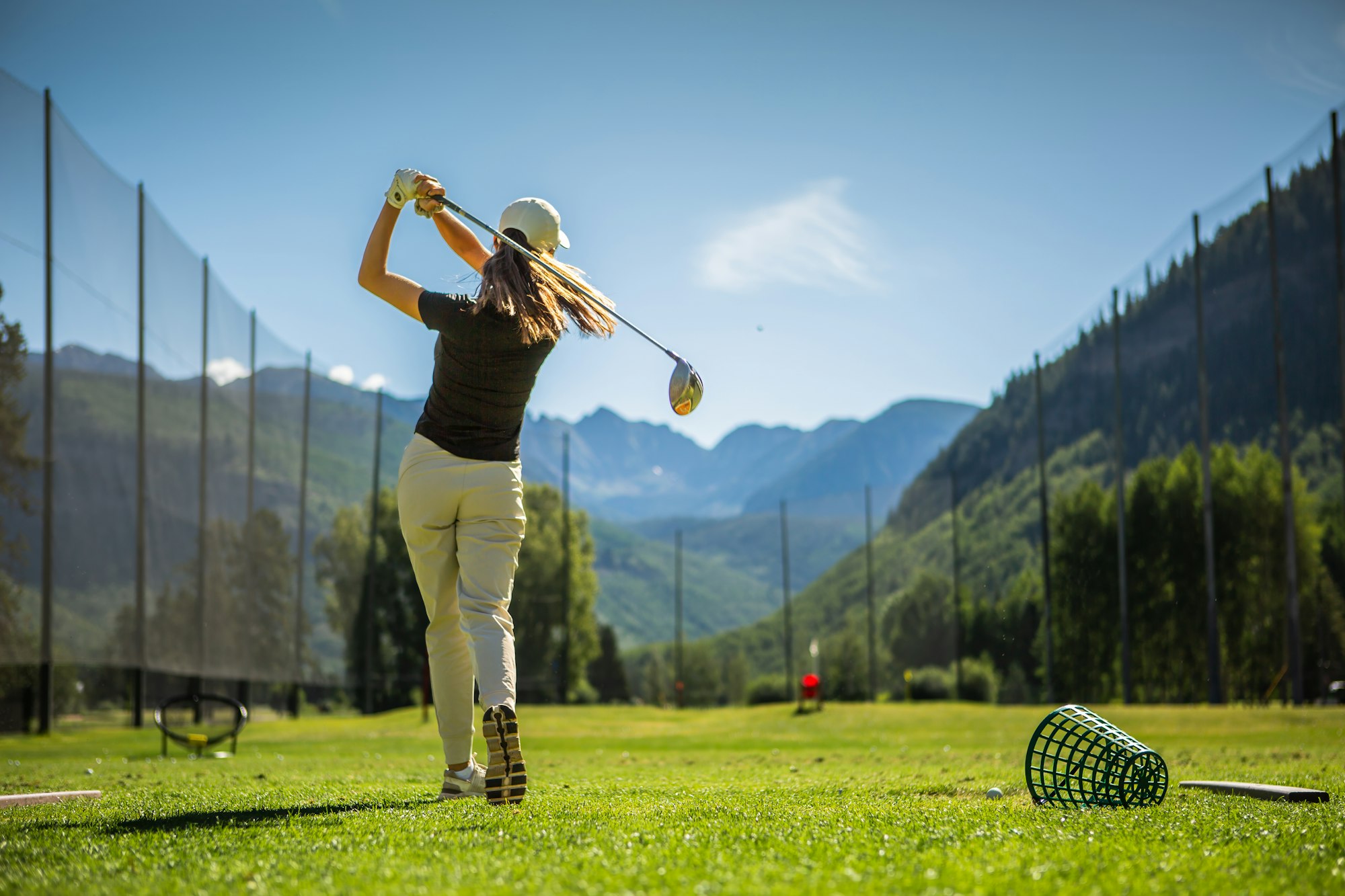 A golfer in mid-swing at a driving range with mountains in the background.