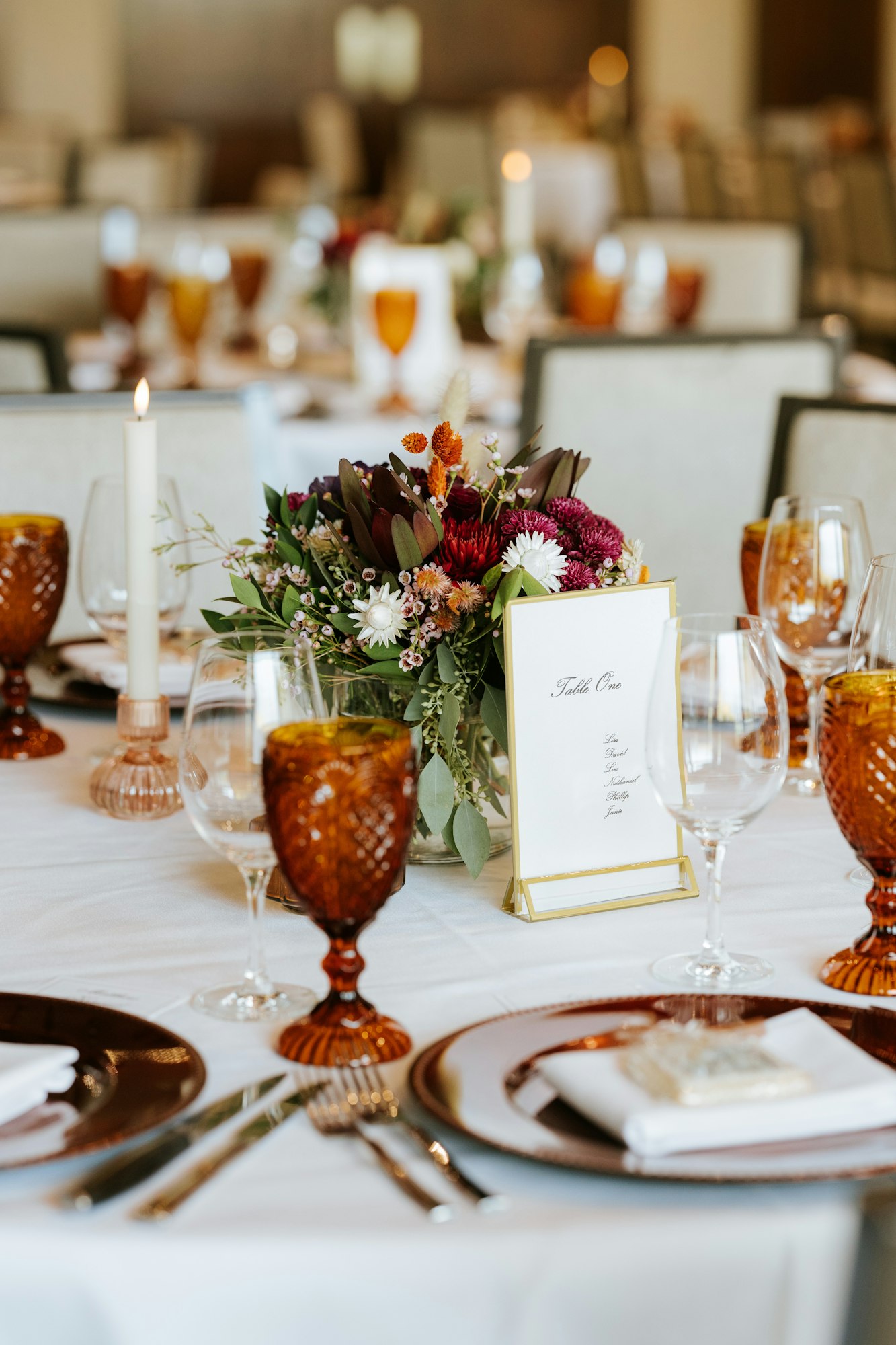 An elegantly set dining table featuring floral centerpieces, amber glassware, a menu card, and a lit candle.