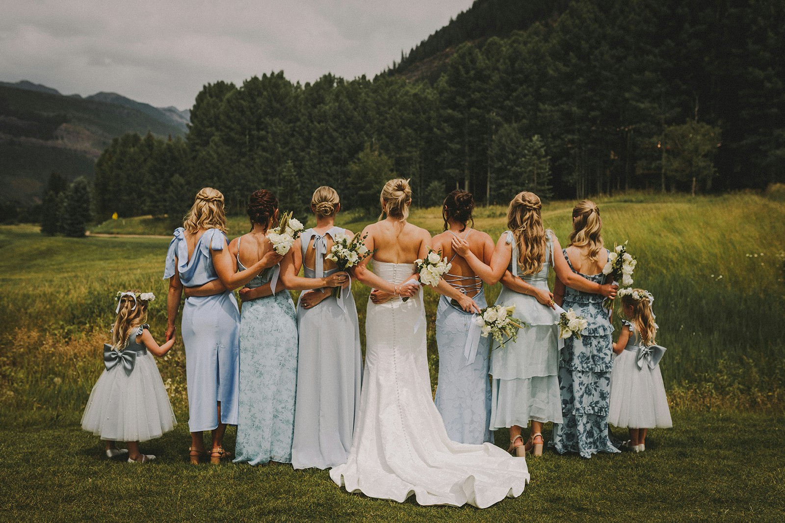 A group of women in blue dresses, including a little girl, posing outdoors, likely for a wedding, with flowers in hand.