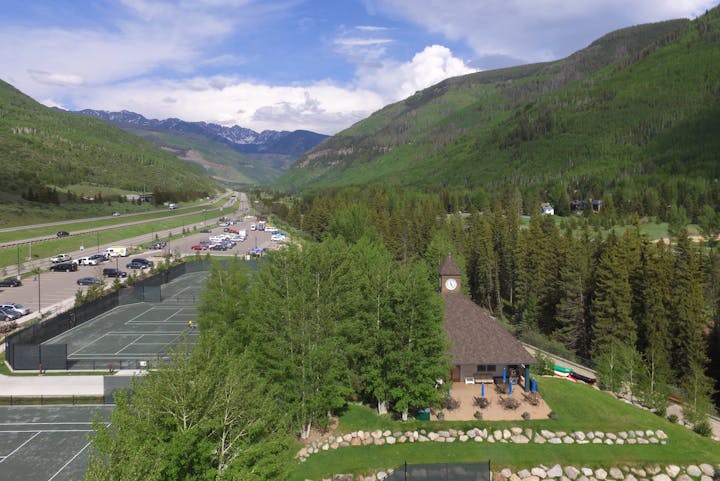 Mountain landscape with tennis courts, a building with a clock, parking lot, highway, and forested hills.