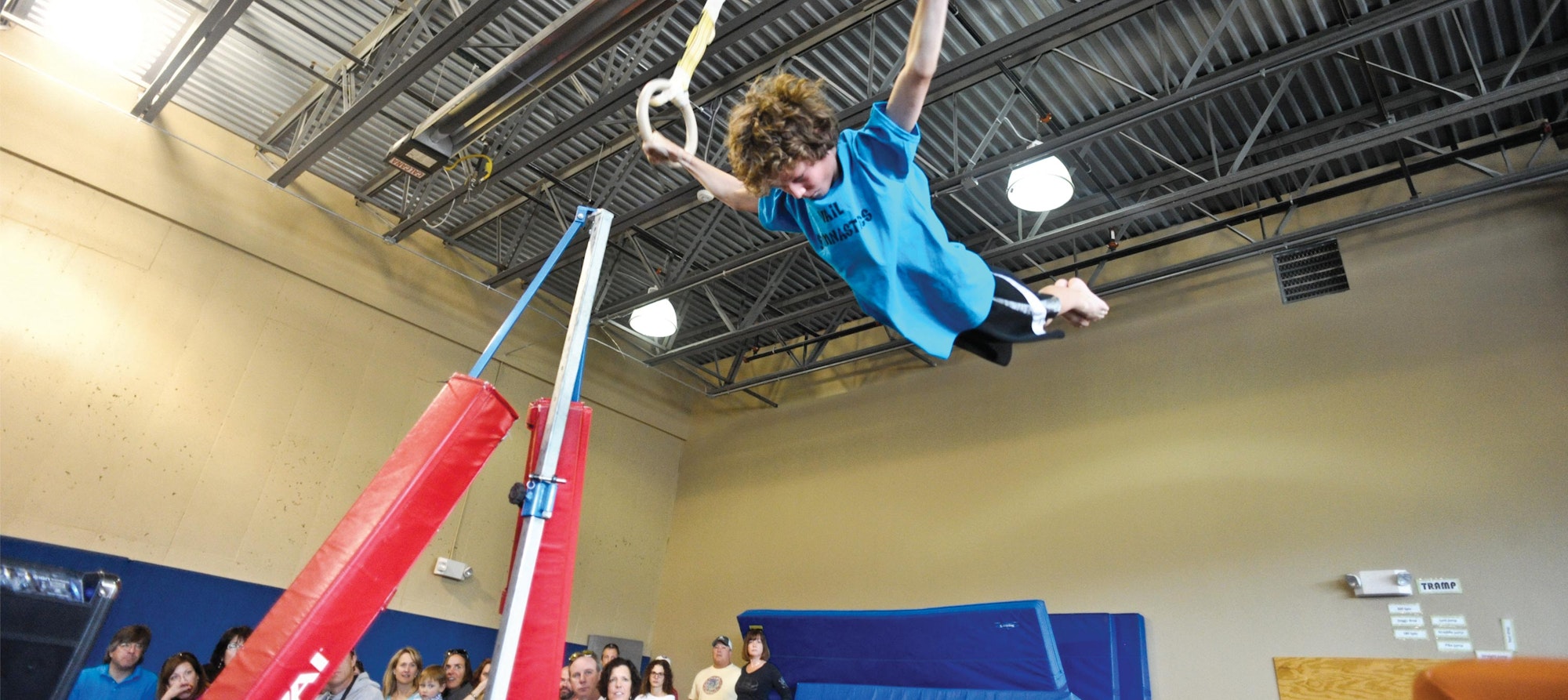 A person performing on gymnastic rings mid-flight, with onlookers in the background.